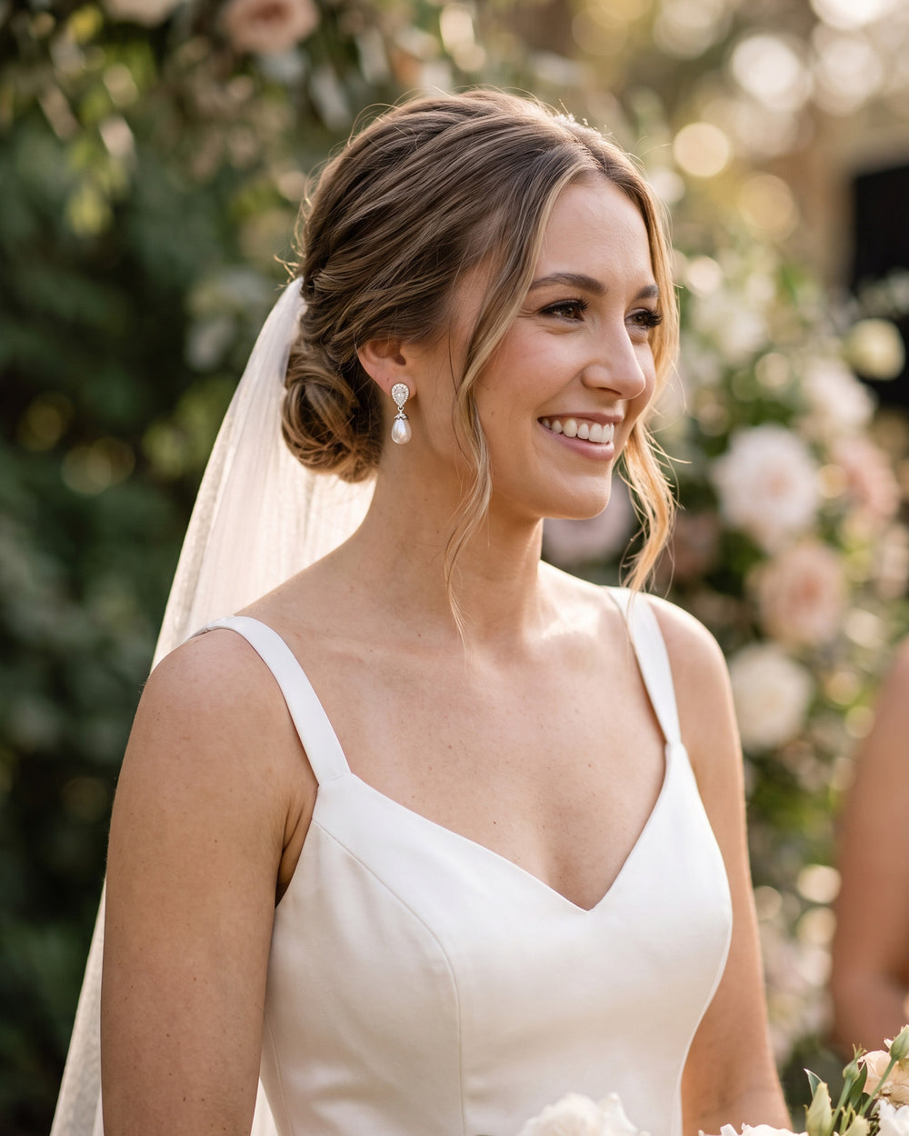 Smiling bride with a veil and low bun wearing Nina pearl drop earrings in a garden