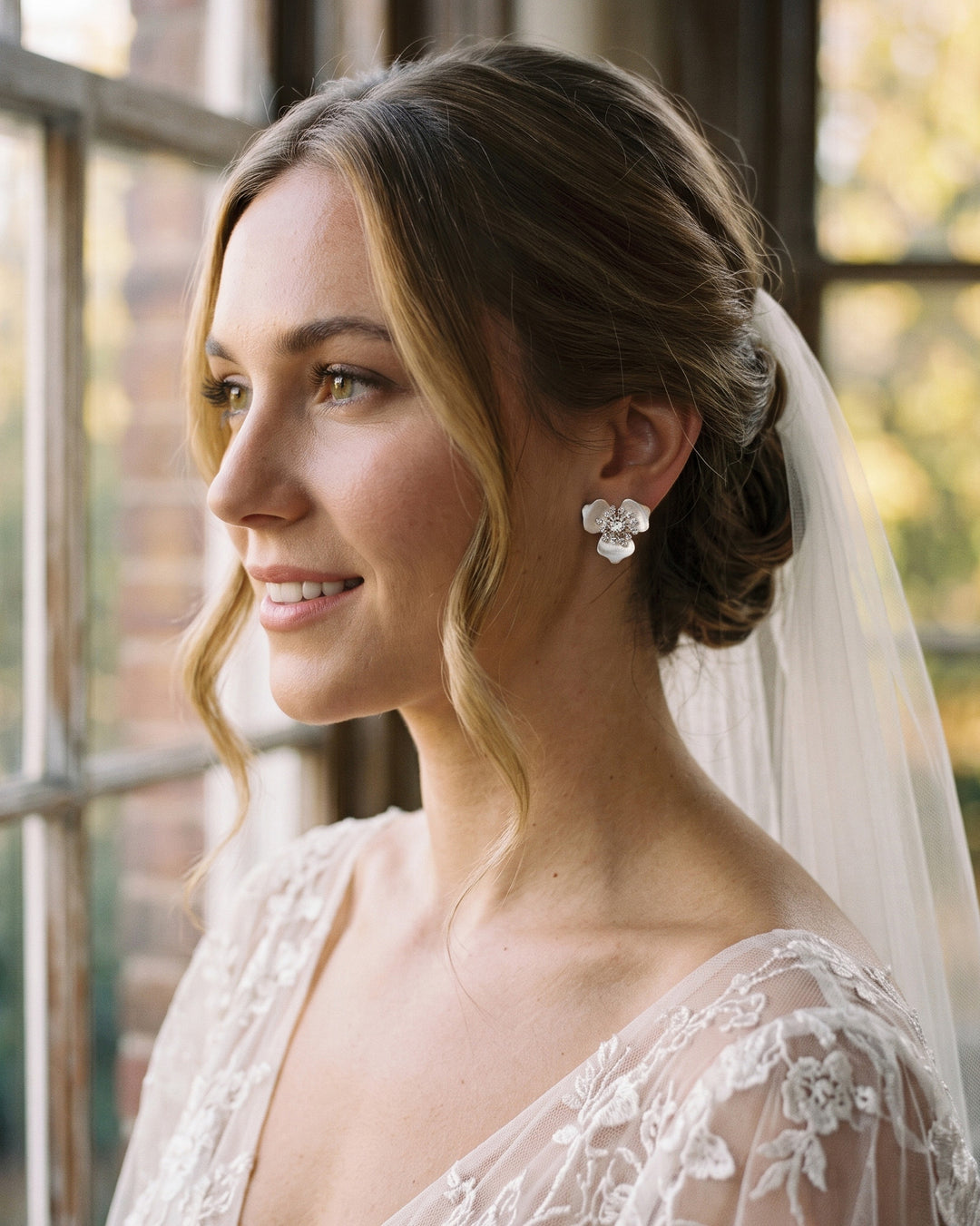 Bride with a veil and low updo wearing Kacie flower studs by a sunlit window