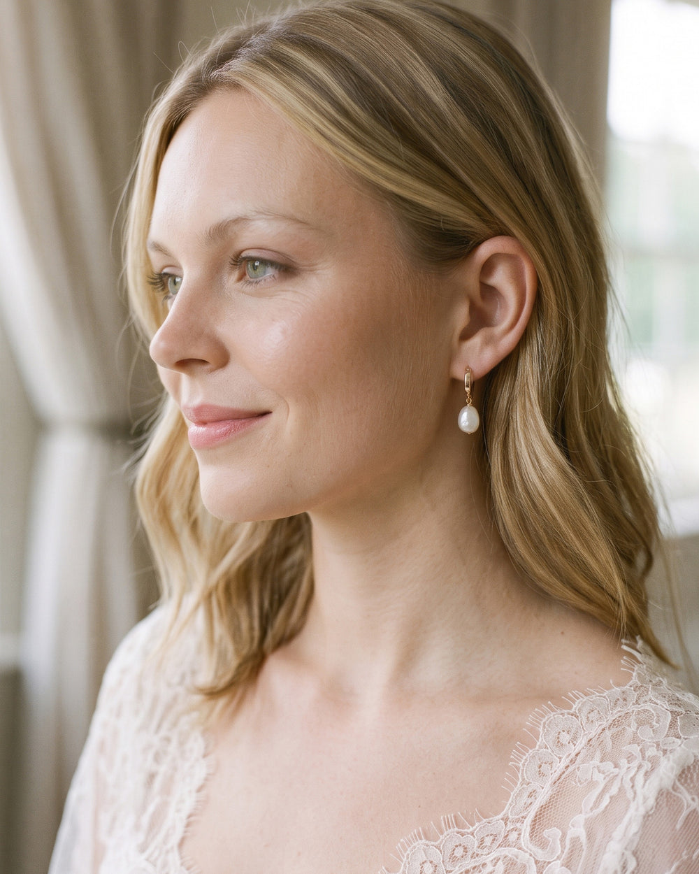 Bride in a lace gown wearing Bali pearl drop huggies by a softly lit window