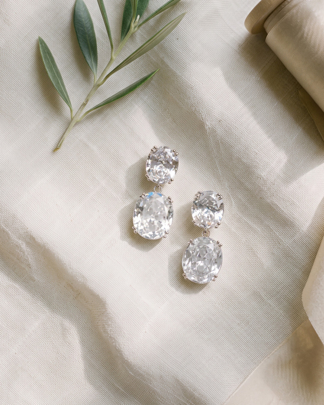 Silver Rosina earrings on ivory linen with an olive sprig, showing the double oval drop