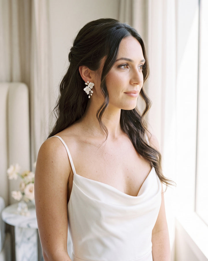 Bride in a cowl-neck slip dress by a window wearing Kara floral wedding earrings
