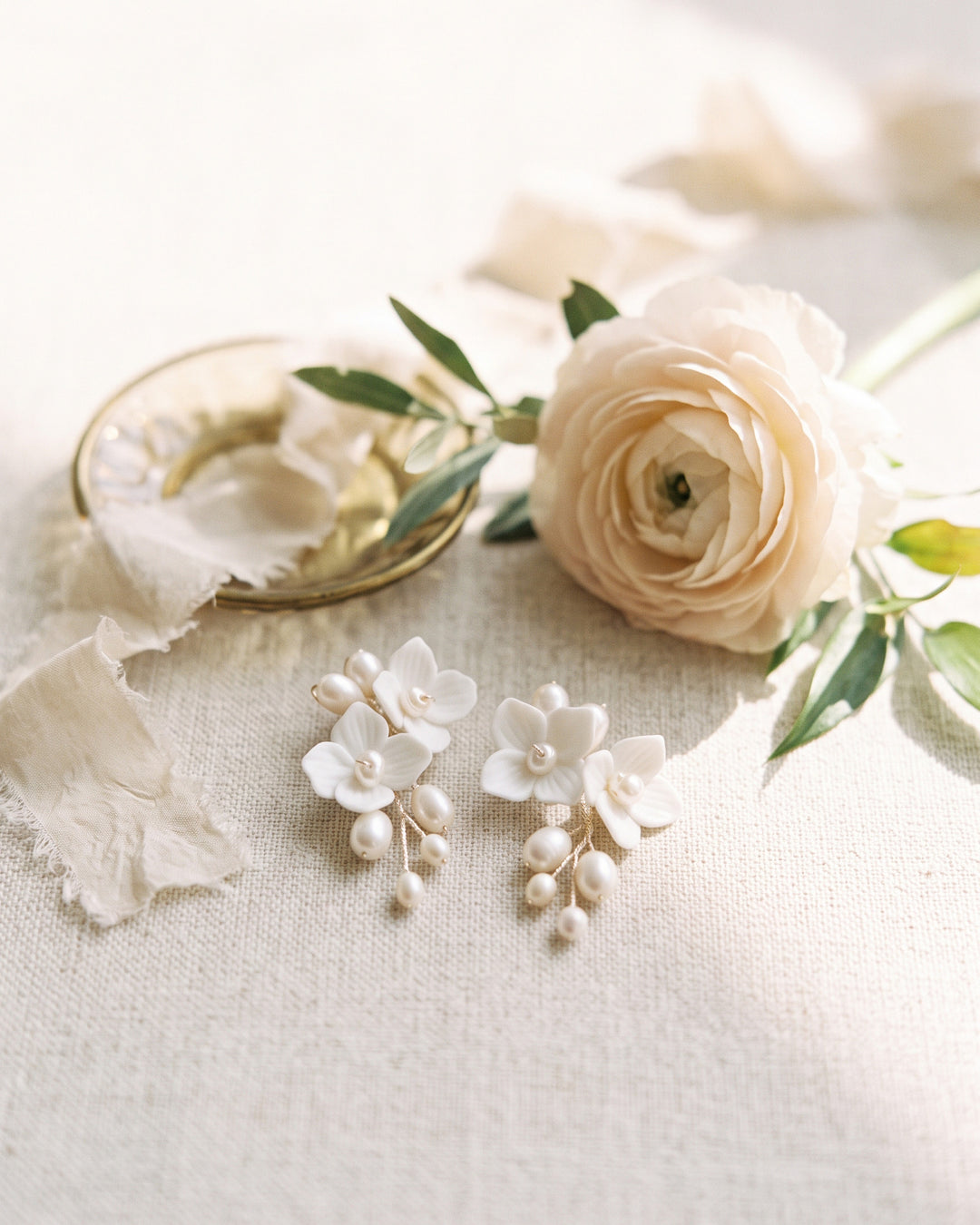 Kara pearl and flower earrings on ivory linen beside a pale ranunculus