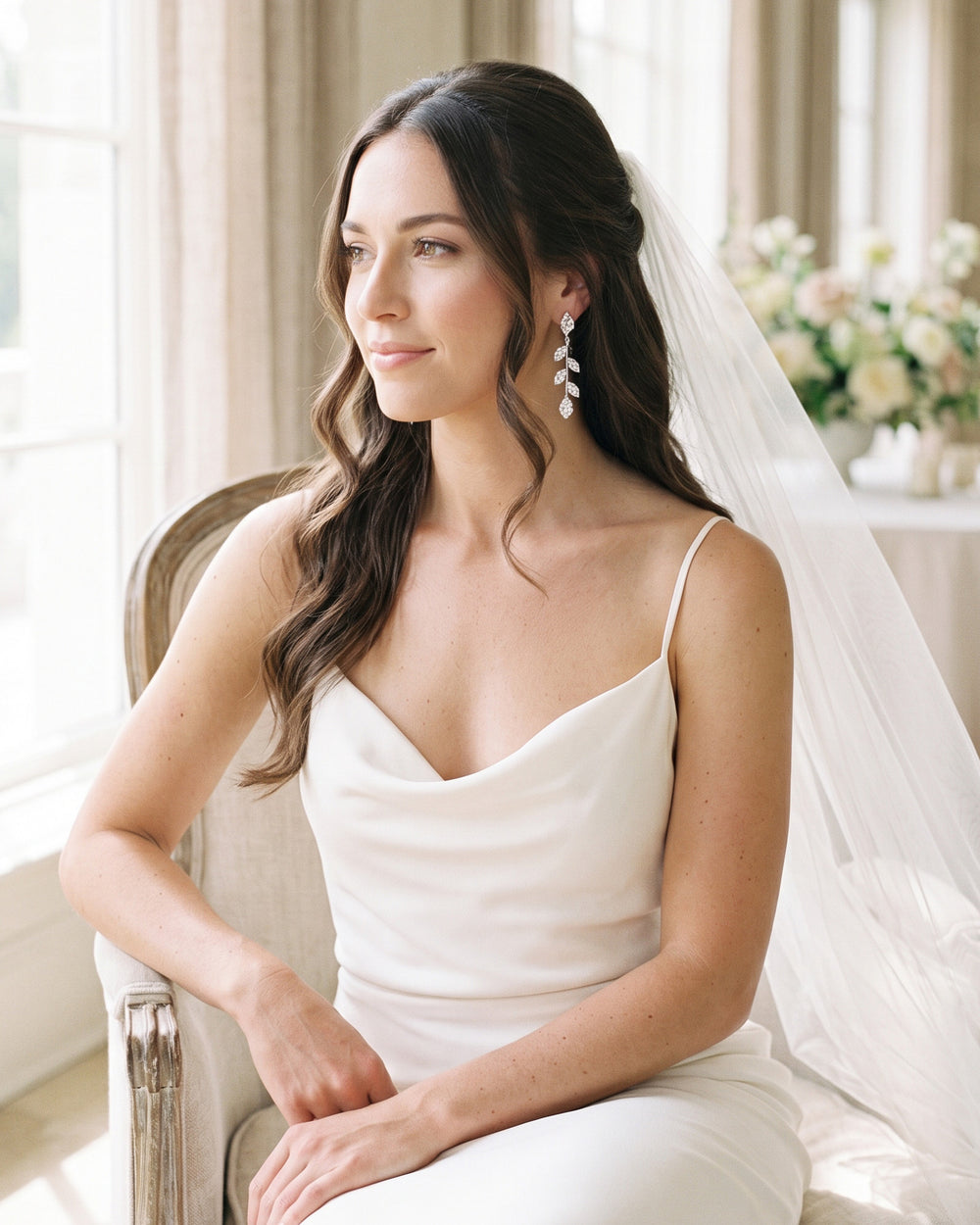 Bride seated by a window in a veil and slip dress wearing Fleur leaf vine earrings