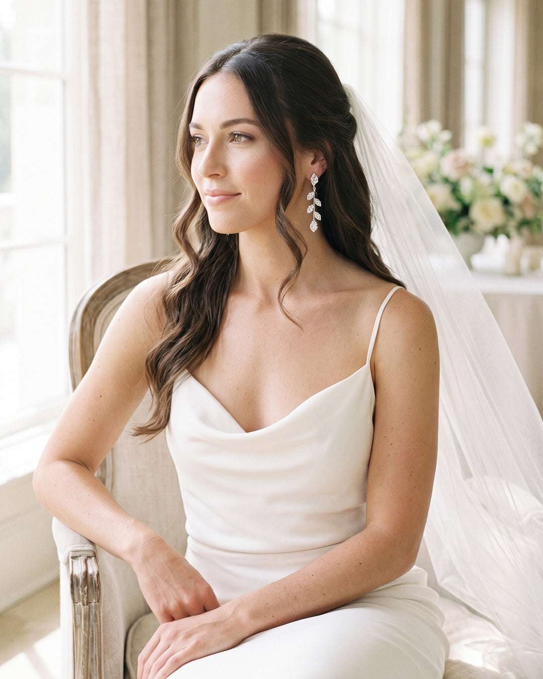 Bride seated by a window in a veil and slip dress wearing Fleur leaf vine earrings