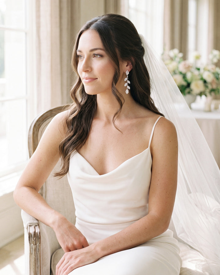 Bride seated by a window in a veil and slip dress wearing Fleur leaf vine earrings