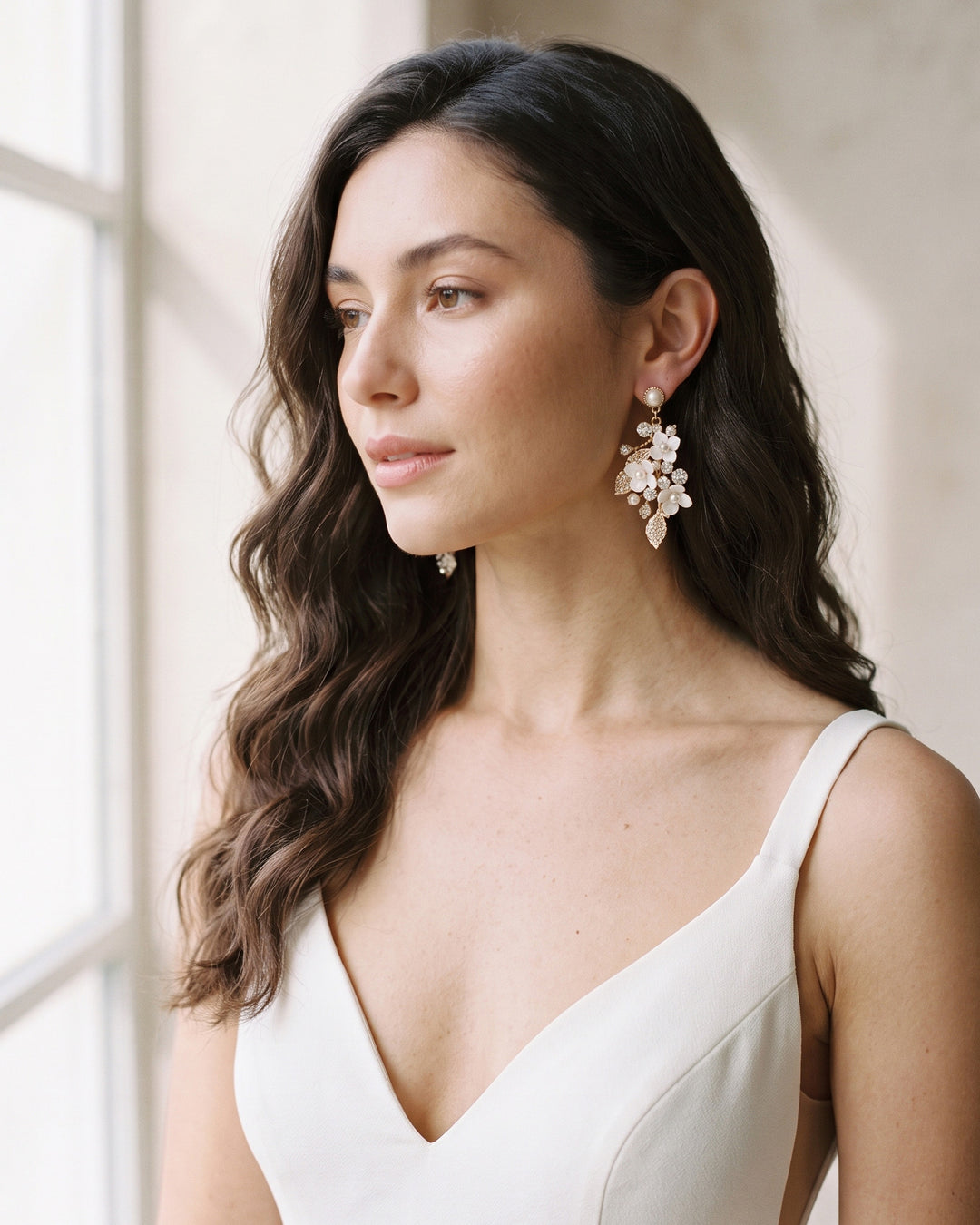 Bride in a V-neck gown wearing gold Lani floral statement earrings by a bright window