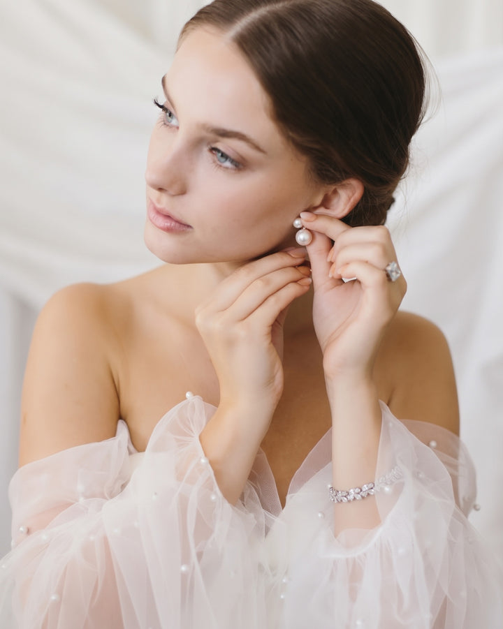 Bride fastening the Nadine pearl earrings while wearing a sheer off-the-shoulder gown