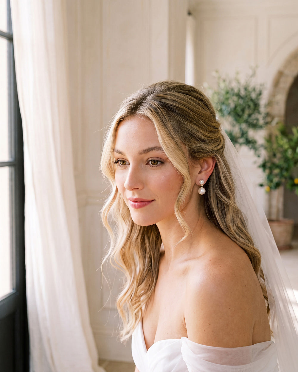 Bride with soft blonde waves and a veil by a window wearing the Nadine pearl wedding earrings