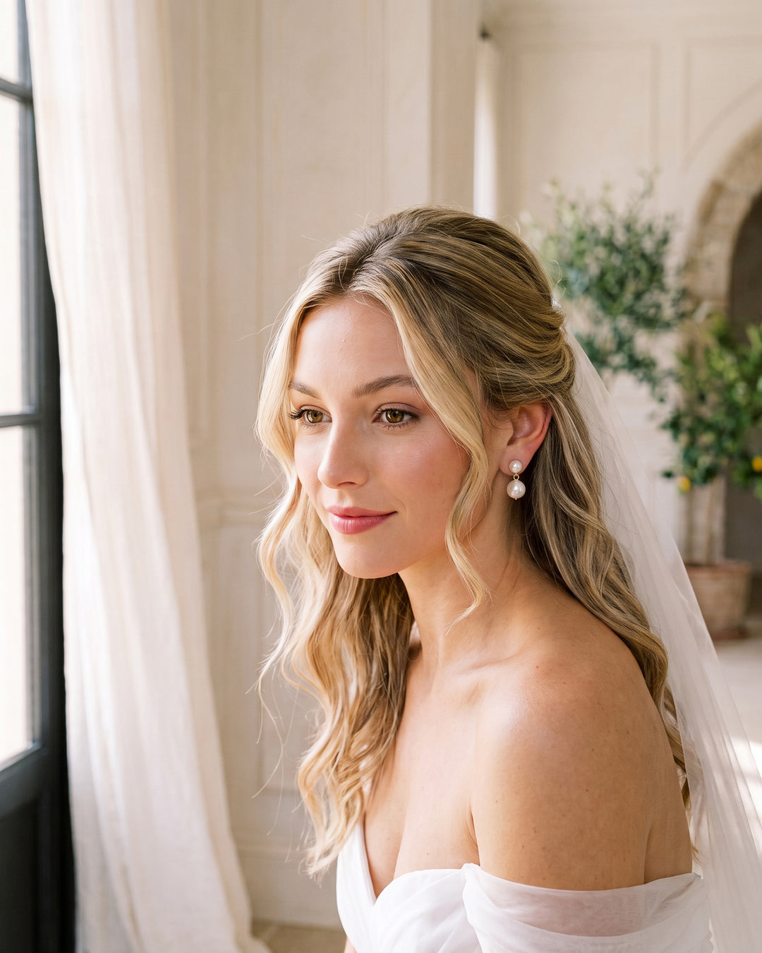 Bride with soft blonde waves and a veil by a window wearing the Nadine pearl wedding earrings