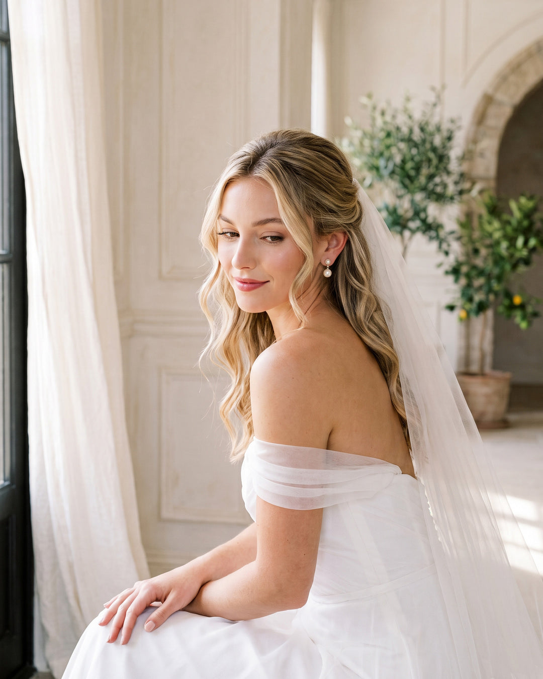 Seated bride in an off-the-shoulder gown and veil wearing the Nadine pearl earrings