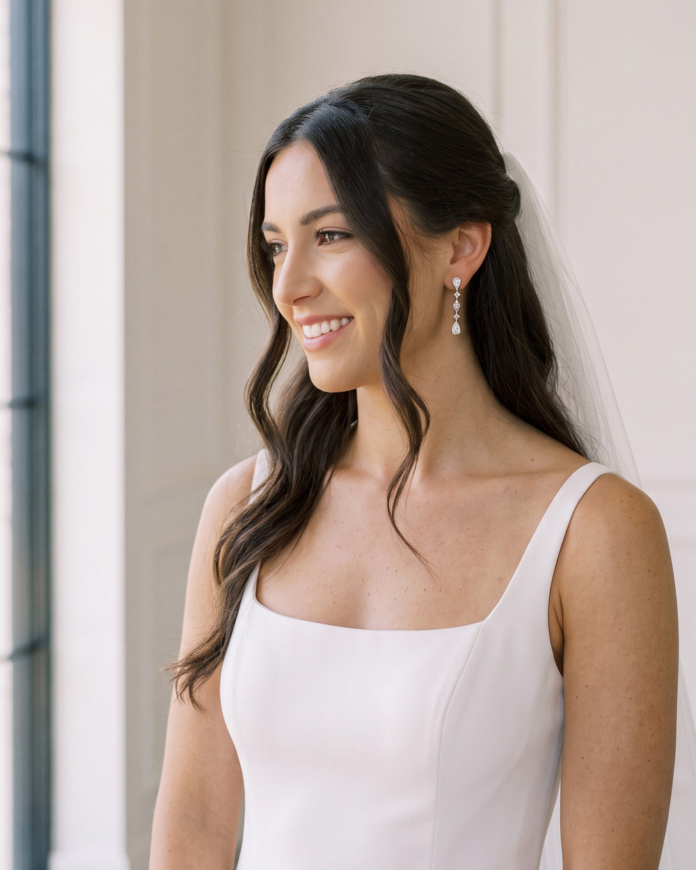 Smiling brunette bride with half-up waves and a veil wearing the Gracie wedding earrings