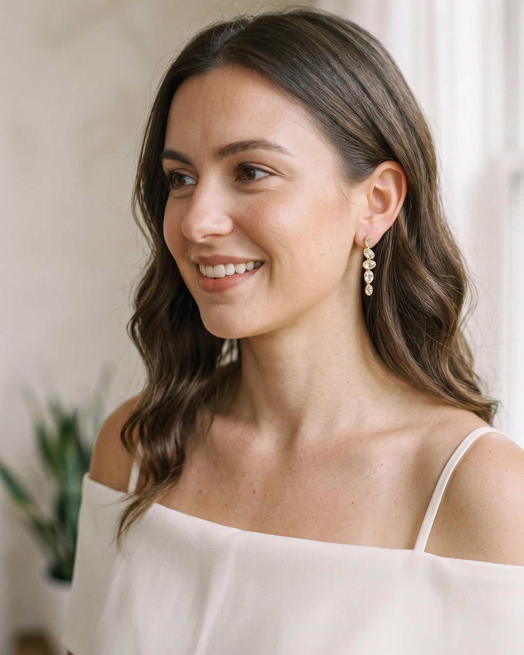 Smiling woman with long wavy hair wearing the Lisbon gold drop earrings in a bright room