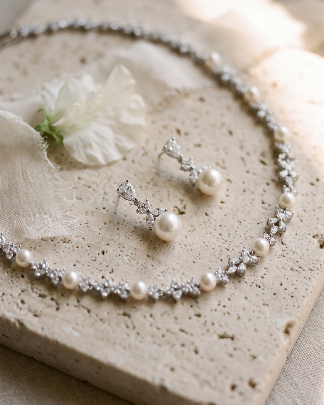 Kelsey pearl necklace and drop earrings laid on travertine stone with a white flower