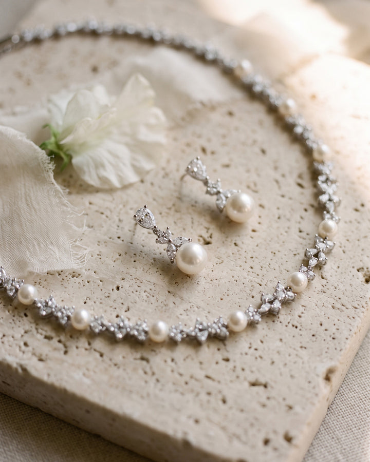 Kelsey pearl necklace and drop earrings laid on travertine stone with a white flower